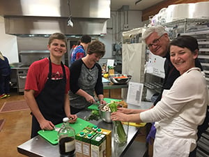 In a community kitchen near downtown Eugene, 14-year-old Eli Panero chops garlic. A lot of garlic. His eyes periodically scan the the counter in front of him for the ingredients he needs to create the recipe, Chicken with Sun-Dried Tomato and Olive Pesto. “I’m really interested in learning how to cook,” he says. “I’d never done it until I came here.”  Behind him, Nicolas and Henry, two 16-year-old students from South Eugene High School, are donning aprons and asking kitchen manager Cody Fuqua what task they should start on first.  This is a typical Monday afternoon at Positive Community Kitchen (PCK), a donation-supported program where teen and adult volunteers come together, alongside trained chefs, to prepare organic meals for people fighting life-threatening illnesses. Many of those free meals have been made for patients of Willamette Valley Cancer Institute.  This week, the volunteers are preparing soups, salads and entrees for 13 families.  “I’d enjoy this no matter who we were cooking for,” Henry says. “But knowing our meals are going to people who are dealing with something so tough, makes it even more meaningful.”     An emotional delivery After the meals are prepared, volunteers, affectionately called “delivery angels,” take them to families’ homes, where they are met with gratefulness, and often tears, for their showing of support. PCK program coordinator Andrea Wolf knows what it feels like to receive such a gift. She was diagnosed with Primary CNS Non Hodgkin’s Lymphoma in January 2013. After undergoing surgery to remove a tumor in her brain, she endured six months of intense chemotherapy. During which, friends and family brought meals to her and her husband, which Andrea says not only nourished her body, but also her soul.  “It’s difficult to shop and cook when you’re not feeling well. Knowing that there was a group of people cooking for me and sending their love through those meals was incredible. Going through cancer is a very scary time and that simple gesture makes a world of difference.”  Following her treatment, Andrea wanted to offer something similar to others.  When she crossed paths with Positive Community Kitchen, and learned of the impactful work they were doing, she immediately joined the team.     A healthy formula To create well-balanced, nutritionally rich meals, PCK embraces the American Institute of Cancer Research’s New American Plate concept, with 2/3 of each meal consisting of vegetables, legumes and whole grains. In addition, PCK focuses on foods that are locally grown. Those who receive the meals get the nutrients needed to aid healing, while teen volunteers learn how to prepare foods in new ways.  “They’re learning that cooking doesn’t have to be complicated, and that good healthy food can taste great,” Cody says. “And, at the same time, they learn that these meals fulfill an important need in the community.”  “We regularly receive thank you cards from families, telling us how overwhelmed they are by the support,” Andrea says. “We all feel fortunate to be a part of that.”  Next month, PCK will expand its program, preparing meals two days a week to serve more people. The program is currently recruiting teen and adult volunteers, as well as people who would like to deliver the meals.  To volunteer or make a donation to support the program, click here.